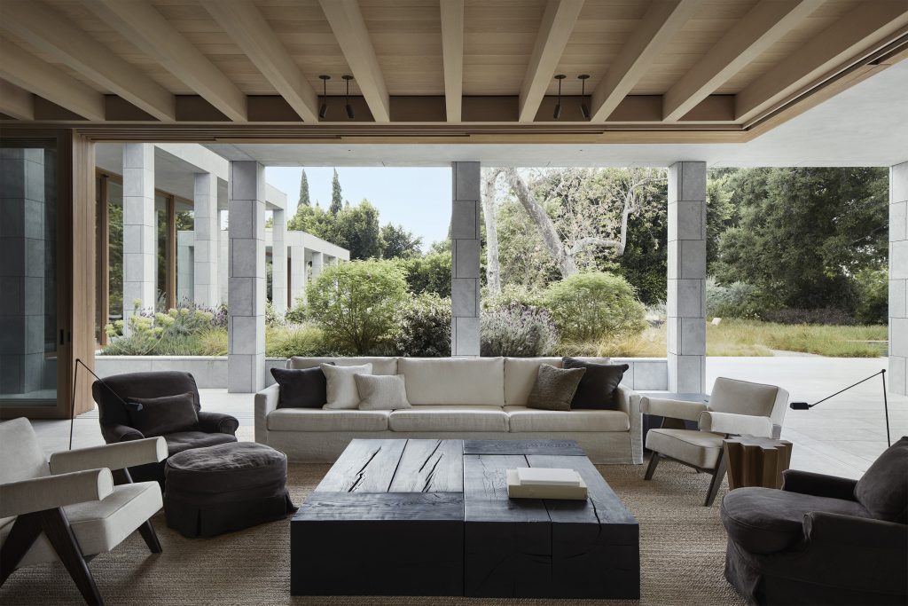 a mid-century modern-inspired living room with mixed fabric and dark wooden lounge chairs centered around a massive, dark charred-wood block coffee table, opening entirely to an architectural stone garden. ELLE Decor A-List