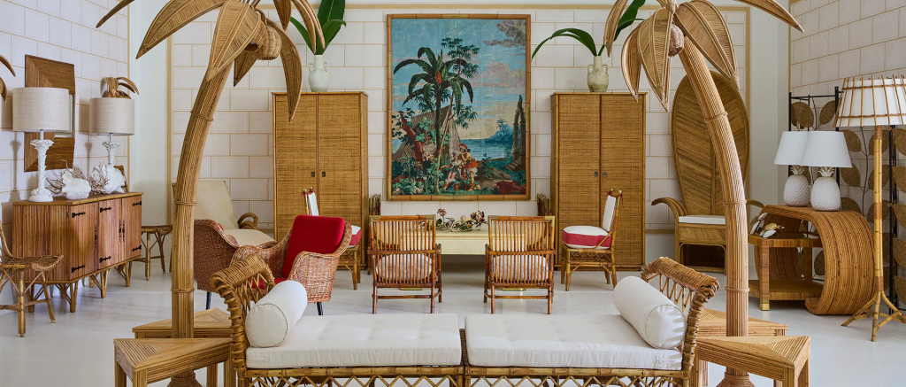 A room decorated with tropical rattan furniture, two palm tree sculptures, and a large decorative tapestry. ELLE Decor A-List