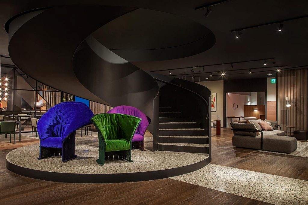 A grand interior space featuring a large, dark, sculptural spiral staircase as the centerpiece. Below and on the landing, unique high-backed, tufted velvet armchairs in cobalt blue, emerald green, and magenta are arranged as statement pieces. The floor is dark wood with textured light-colored stone paths. In the background, a modular sofa and other luxury home furniture are visible. Warm, directional spot lighting illuminates the area. luxury showrooms