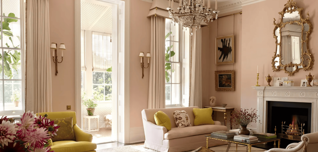 A sophisticated living room with soft pink walls, a classic fireplace, ornate gold mirror, and elegant furnishings. ELLE Decor A-List