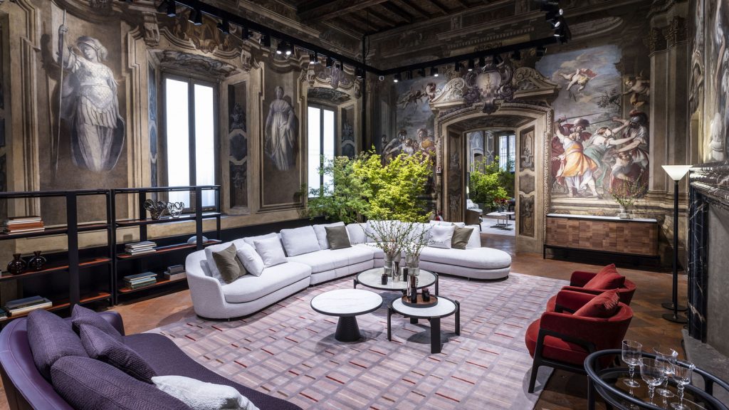An interior view of a majestic living room in a historical palace, adorned with detailed wall frescoes and classical architectural elements. A curved white sectional sofa sits at the center, surrounded by two red velvet armchairs and multi-level coffee tables, luxury showrooms.