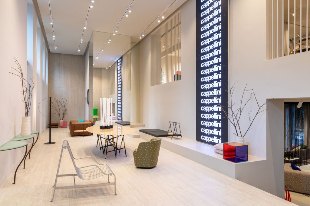 A sophisticated, high-end furniture showroom with polished pale limestone flooring. The space is minimalist, featuring curved metal furniture like a console table with tall candles and a modern armchair with a grid pattern. A large black display panel lists the brand name "Cappellini" in repeating white text, acting as a dynamic visual wall. A modular leather sofa is in the background, with dry branches providing organic decor. luxury showrooms