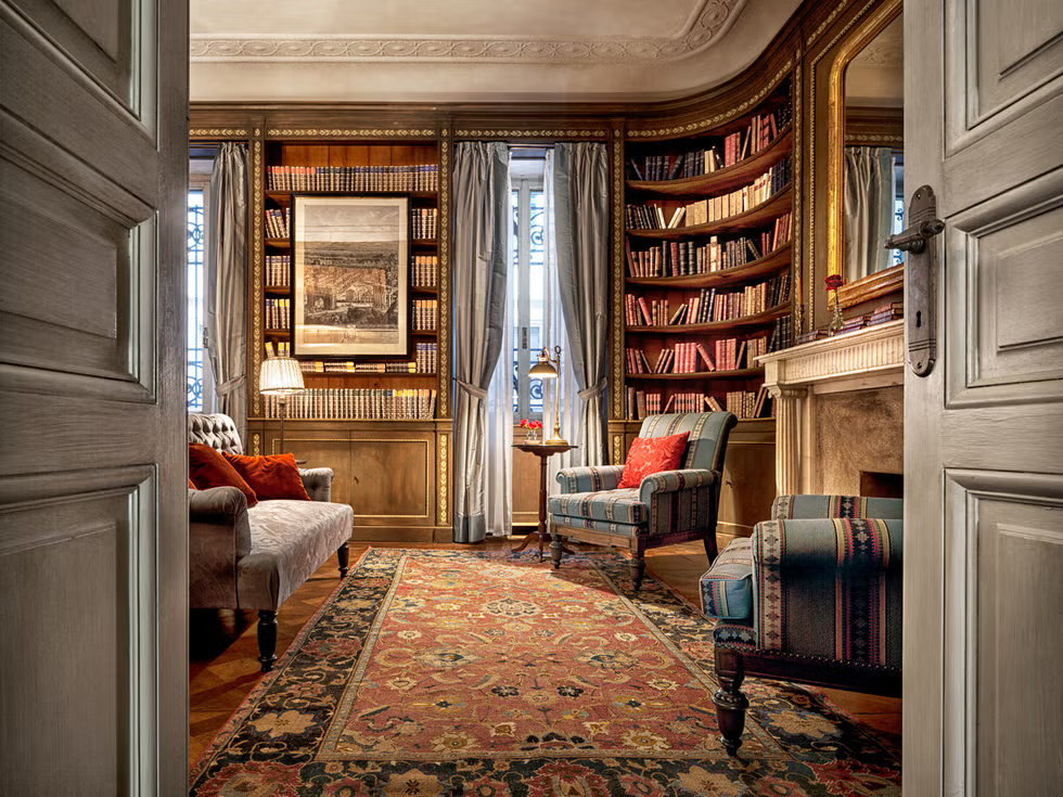 an intimate, classic wood-paneled library viewed through French doors, featuring curved bookshelves, a small fireplace, a vintage patterned rug, and upholstered armchairs. ELLE Decor A-List