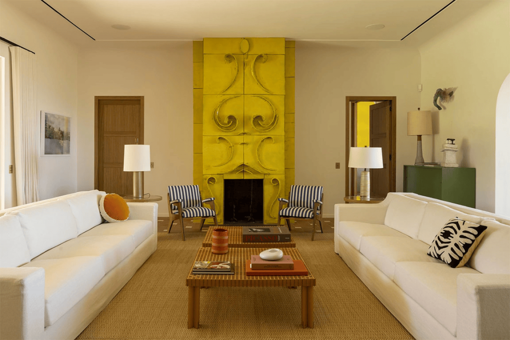 two long white sofas face each other over low wooden coffee tables on a large tan area rug in a modern living room featuring an ornate, sculpted bright-yellow fireplace mantel. ELLE Decor A-List