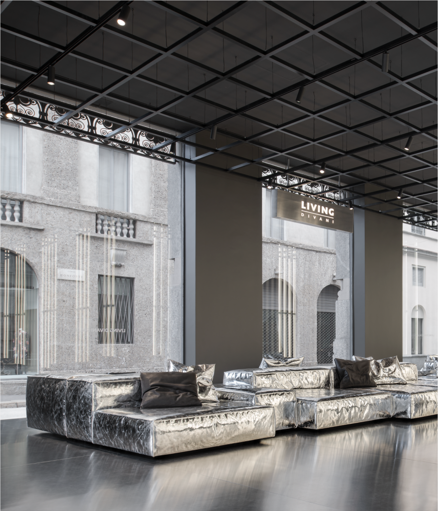 A close-up view through a high-arched glass window overlooking a historical city street, focusing on an interior display. A long, modular sofa is wrapped in shimmering, textured silver metallic fabric with dark cushions, contrasting with the dark industrial ceiling grid, luxury showrooms.
