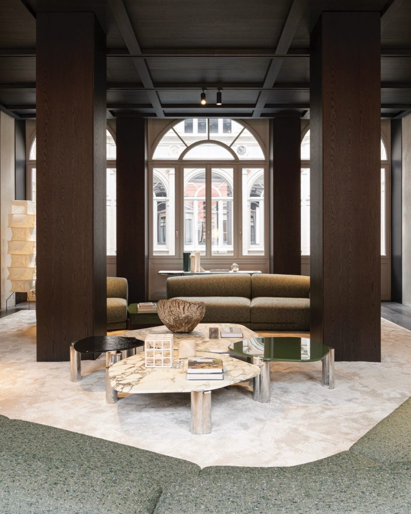 A focused interior view looking through two massive dark wood support columns into a luxury lounge area. A group of multi-level marble and glass-topped coffee tables rests on a patterned rug, facing a plush green sofa in the background, luxury showrooms.