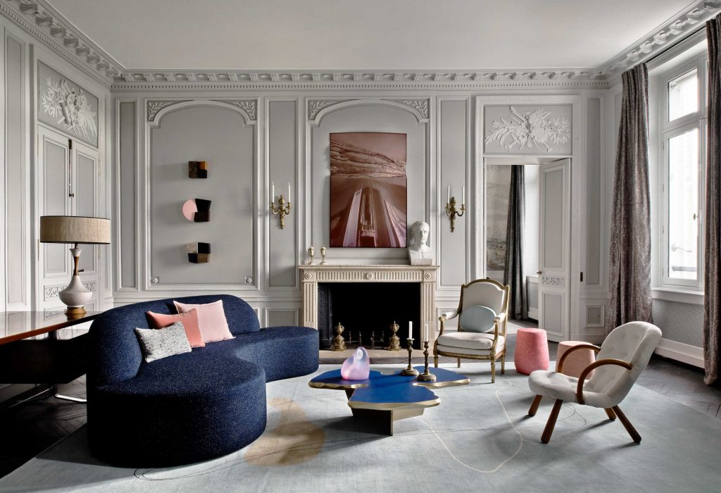 An elegant and sophisticated living room, characteristic of classic French interior design, as presented in the living room. The space features gray walls adorned with detailed moldings, a central fireplace topped by a contemporary artwork and a bust sculpture. The furniture includes a deep blue curved sofa, an organic-shaped coffee table in blue with gold accents, a mix of classic and modern armchairs, and a light-toned rug with subtle patterns. The lighting is soft, and the overall space conveys a luxurious and refined atmosphere.  ELLE Decor A-List