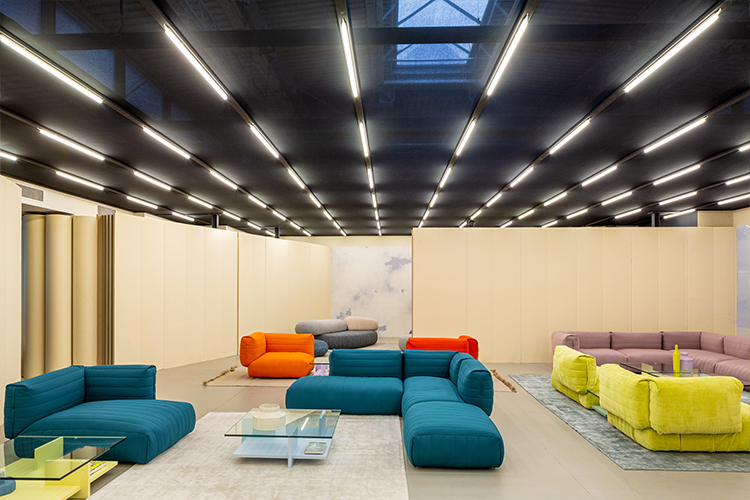 A photograph of a large interior space with a distinctive linear tube lighting grid on a dark ceiling. Modular sofas in teal, orange, and lime green are arranged in small vignettes across the space, bordered by high-standing pale wooden panel dividers, luxury showrooms.