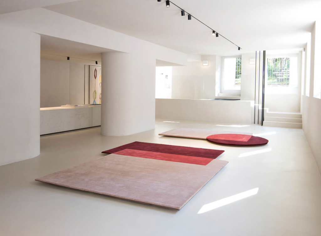 A minimalist, art gallery-like showroom for luxury rugs. The vast space has a smooth concrete floor and clean white walls with rounded columns. Minimalist black track lighting hangs from the ceiling. Multiple large-format rugs, including a rectangular rug with bands of dusty pink and burgundy, and a circular rug with a color-block pattern, are artfully positioned on the floor. Sunlight streams in from large windows, creating dramatic light patterns. luxury showrooms