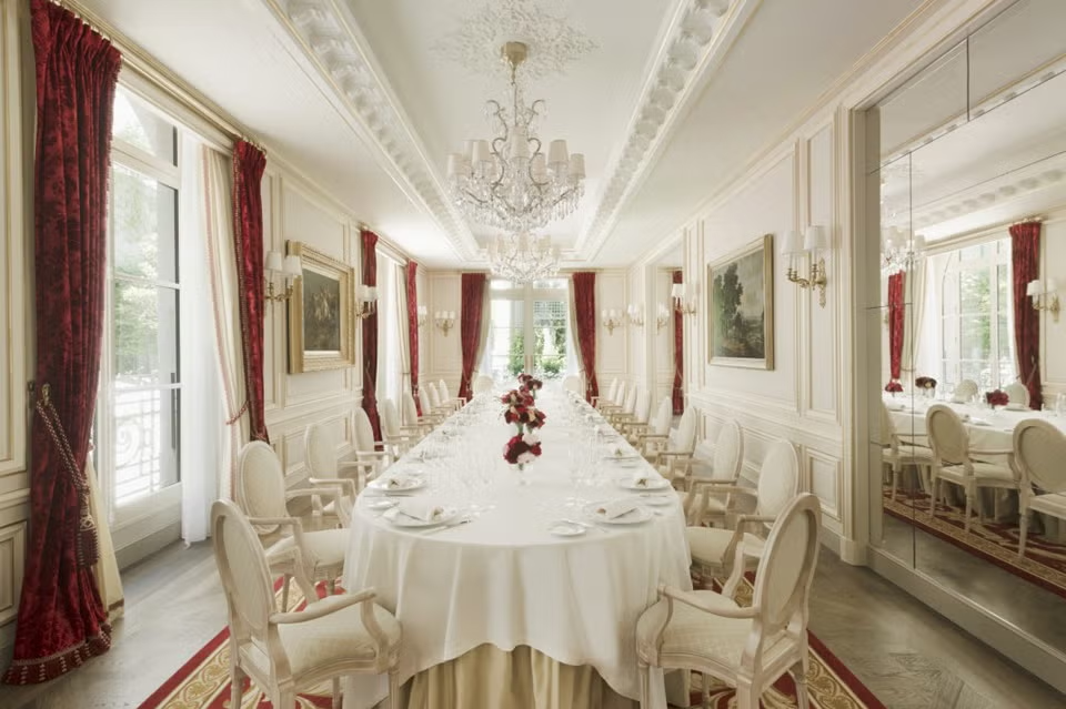 A formal, long dining room with a large table, white chairs, red velvet curtains, and a crystal chandelier. ELLE Decor A-List
