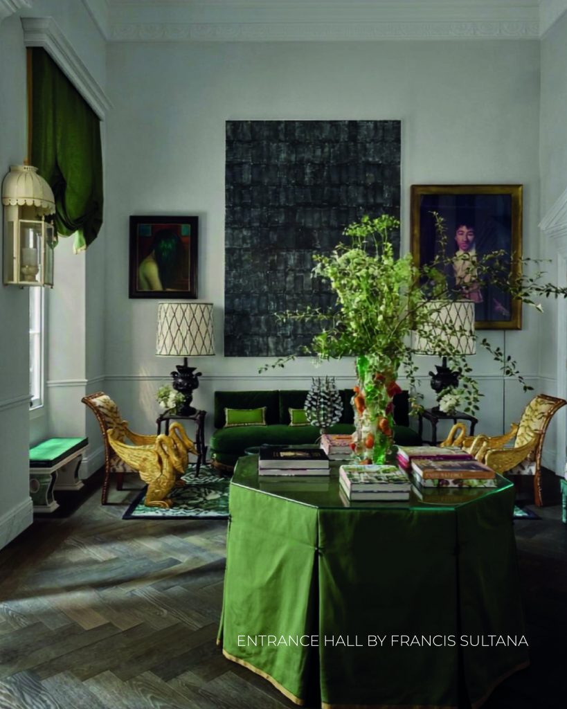 Hall de entrada amplo com decoração aristocrática e elementos de design marcantes. No centro do espaço, destaca-se uma grande mesa octogonal coberta com uma saia de tecido verde-oliva e tampo de vidro, sobre a qual repousam livros de arte e um arranjo alto de ramos verdes e flores. Ao fundo, um sofá de veludo verde-escuro está posicionado entre duas imponentes poltronas douradas esculpidas em forma de cisnes majestosos. Grandes candeeiros de mesa com abajures de padrão geométrico flanqueiam o sofá. Na parede cinzenta clara, destaca-se uma enorme pintura abstrata preta e escura ao centro, ladeada por dois retratos emoldurados. O chão é de parquet de madeira escura em espinha. Na base da imagem, lê-se o texto "ENTRANCE HALL BY FRANCIS SULTANA". WOW!house