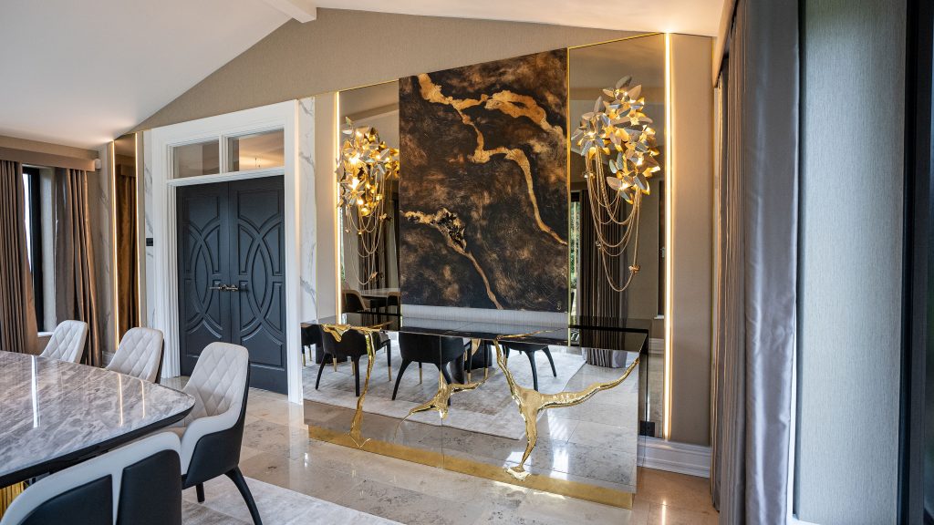 Sophisticated dining room with a glossy mirrored and brass sideboard featuring sculptural gold leg details, a large abstract gold and black artwork, gold branch-style wall sconces, and a marble dining table in the background kunal trehan
