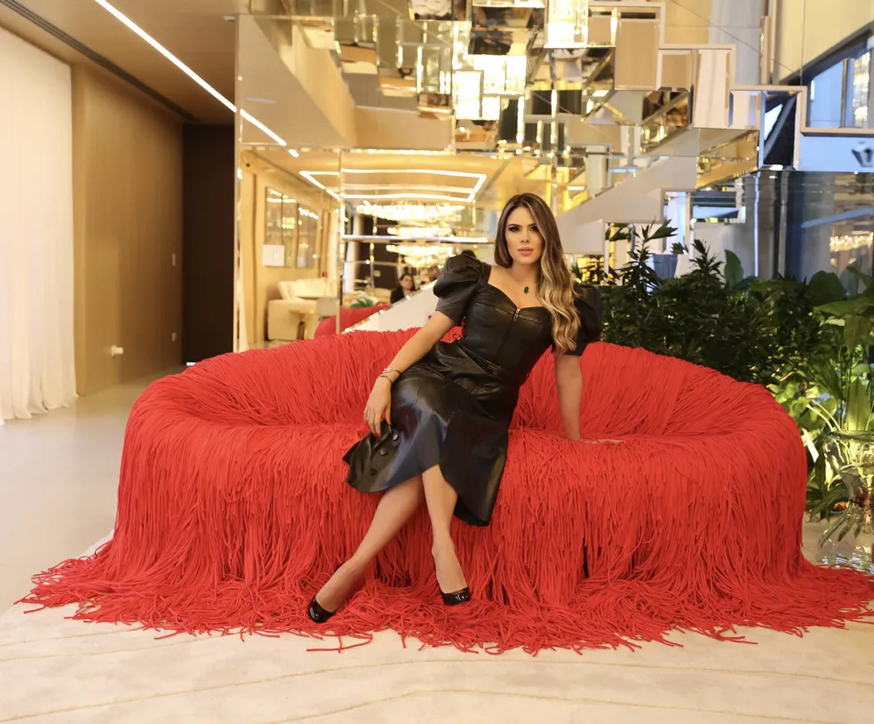 Woman in a black leather dress with puff sleeves sitting on a red fringe sofa in a modern lobby with mirrored ceiling panels and gold accents. showroom de luxo em sao paulo