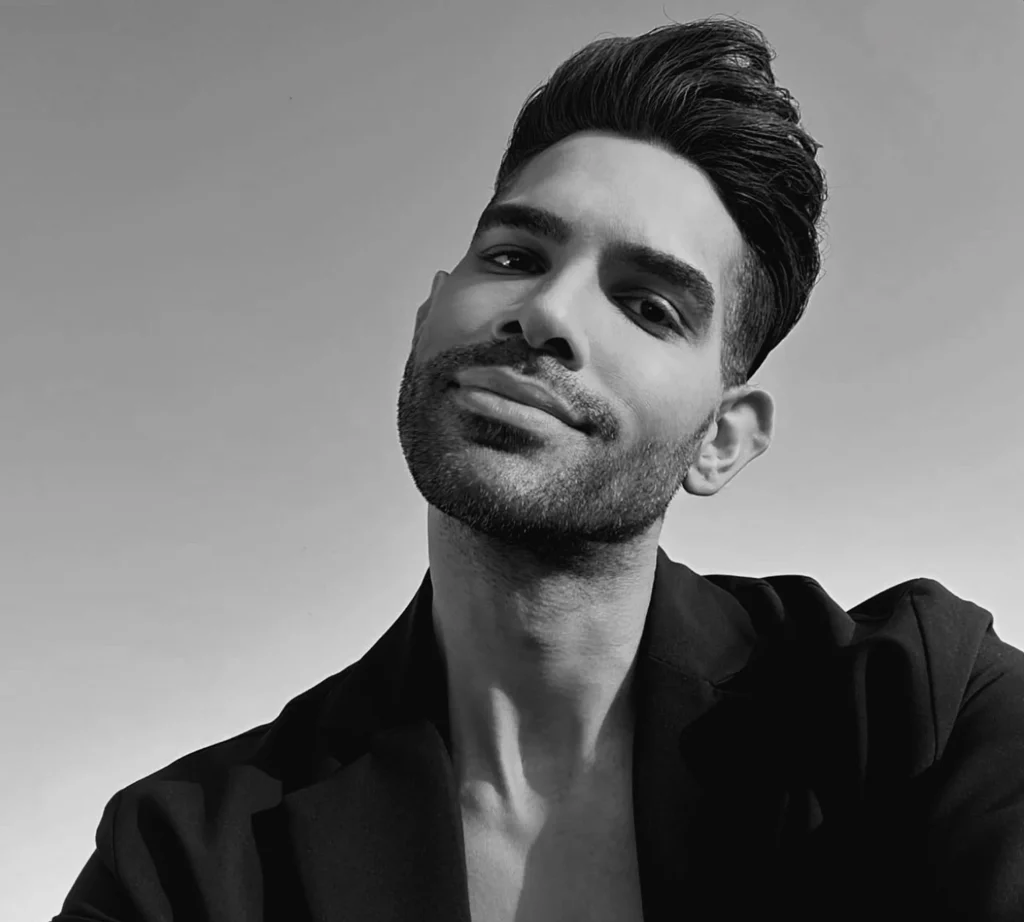 Black and white portrait of a man with styled dark hair and light stubble, wearing an open black blazer, looking down at the camera against a plain background. kunal trehan