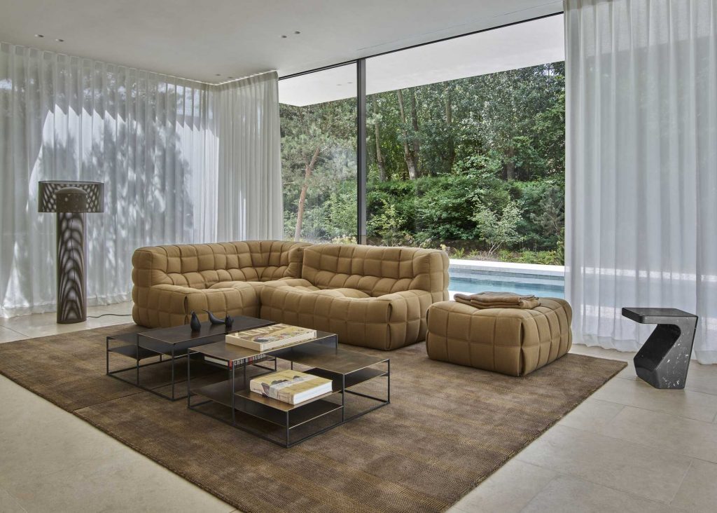 A contemporary and airy living room with a floor-to-ceiling glass wall overlooking a lush green forest and a swimming pool. The focal point is a large, tufted modular corner sofa in a warm beige hue, accompanied by a matching ottoman. Two black stacked geometric coffee tables with bronze accents, a sculptural floor lamp, and a textured brown rug complete the space. luxury furniture
