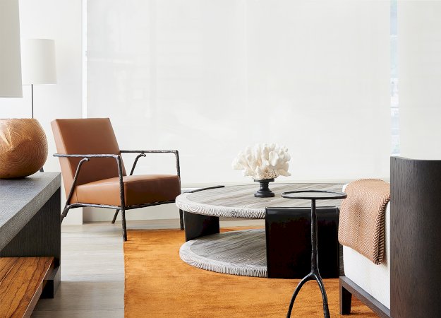An elegant and modern living room with luminous white glass walls. A cognac leather armchair with a sculptural metal frame and wood accents is the highlight, paired with a dark wood sofa featuring white linen upholstery. The coffee table is a round reclaimed wood design with a glass top, displaying a large white coral. A slim black metal side table and a burnt orange area rug add color and texture. luxury furniture