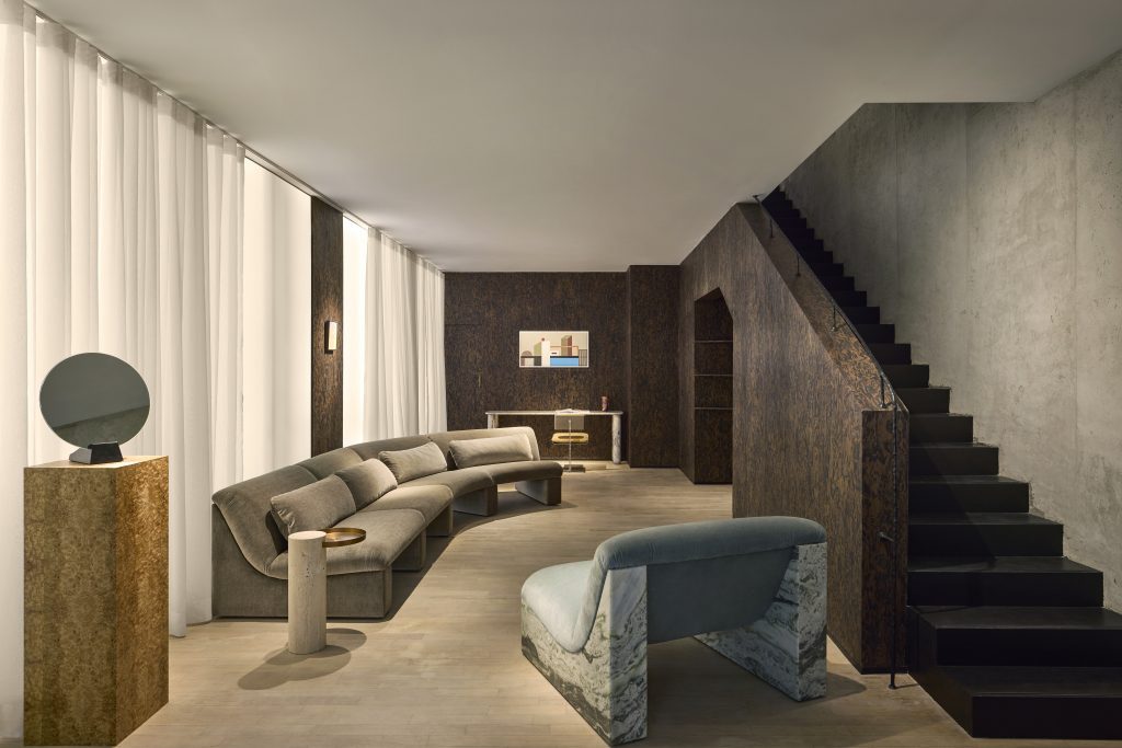 A luxurious and modern living room interior with a warm color palette and earth tones. It features a long, curved grey-taupe velvet sofa, a unique teal velvet armchair with a marble base, and a cylindrical travertine side table. The background is a dark, textured wood wall, complemented by white linen curtain panels. A sleek, modern black staircase with a metal railing ascends along the concrete wall. luxury furniture
