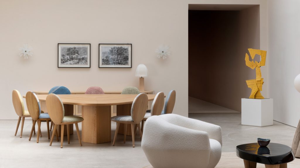 A sophisticated and minimalist dining room with neutral beige walls. A sculptural oak dining table with a rounded triangular top and thick light wood legs is surrounded by upholstered chairs featuring curved wood backs and bouclé fabric seats in varied pastel shades (yellow, blue, pink, green). In the background, an oak console cabinet holds a table lamp and two framed landscape prints. To the right, a vibrant yellow geometric sculpture on a white pedestal and an organically shaped textured white bouclé armchair. luxury furniture