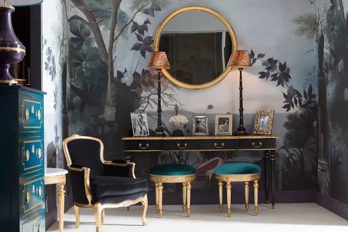 A luxurious and classic boudoir corner, transformed with a dramatic grey and black forest wallpaper mural. A sleek black vanity table with brass hardware and a marble top is centered under a large, gold-framed circular mirror. Two teal bouclé stools with ornate gold legs match a gold-framed black Louis XV style armchair. The floor is light carpeting. luxury furniture