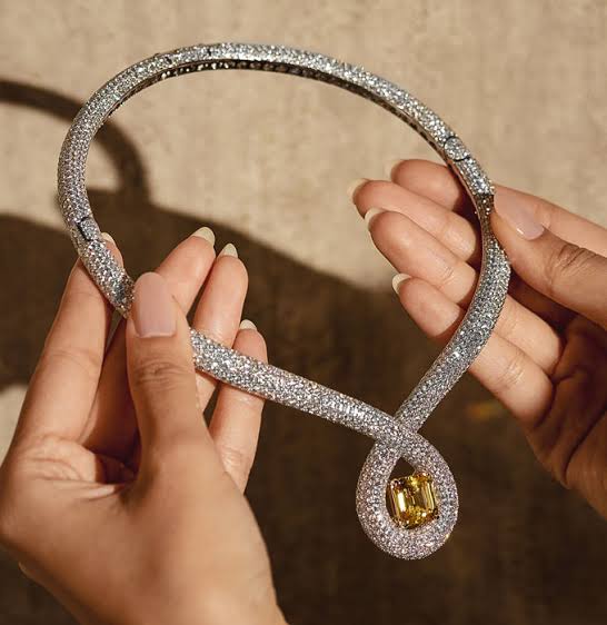 Hands holding a sparkling diamond necklace with a central yellow gemstone, luxury brand