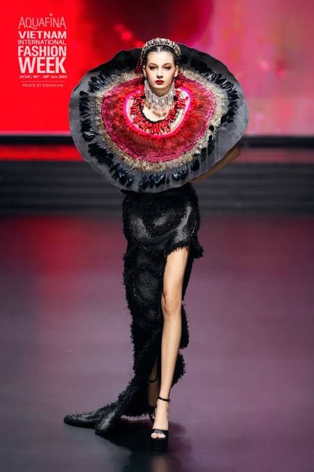 Model walking on a runway wearing an elaborate black dress and a large colorful feathered collar, luxury brand