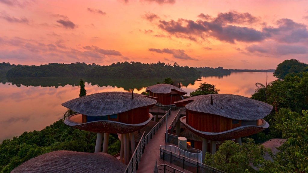 Aerial view of modern round treehouse villas connected by wooden walkways overlooking a serene lake at sunset, luxury brand
