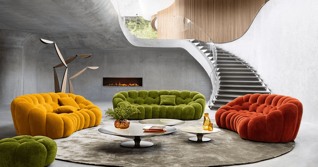A luxurious and modern living room interior with a warm color palette and earth tones. It features a long, curved grey-taupe velvet sofa, a unique teal velvet armchair with a marble base, and a cylindrical travertine side table. The background is a dark, textured wood wall, complemented by white linen curtain panels. A sleek, modern black staircase with a metal railing ascends along the concrete wall. luxury furniture