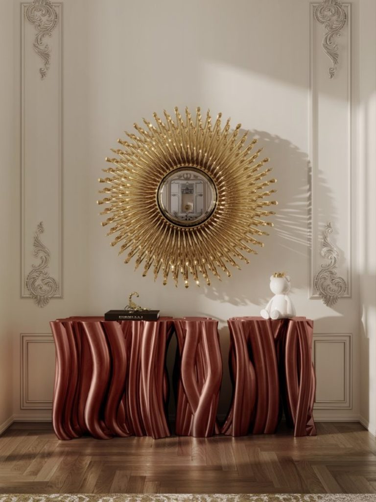 Elegant entryway featuring Boca do Lobo’s golden Goddess Mirror above a sculptural burgundy console with classic wall mouldings.