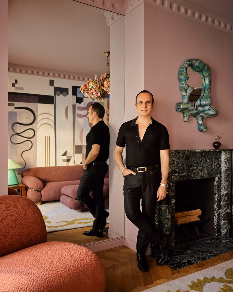 Greg Natale standing beside a mirrored console in a mauve-pink interior with abstract mural and green marble fireplace