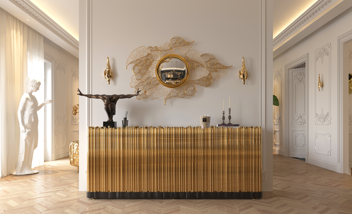 Luxurious classic interior with cream walls, ornate mouldings and herringbone wood flooring, featuring a gold fluted sideboard topped with a dark sculptural figure, candles and decor, beneath an intricate gold filigree wall mirror with matching gold sconces.
