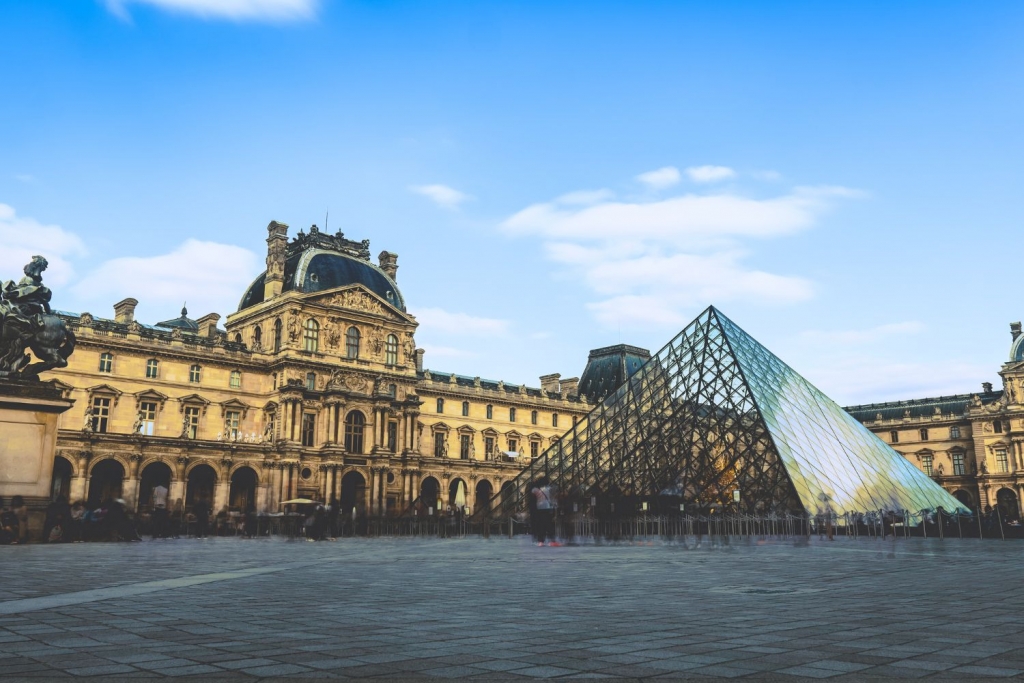 Maison- View of the Louvre Museum in Paris featuring the iconic glass pyramid entrance under a blue sky with scattered clouds