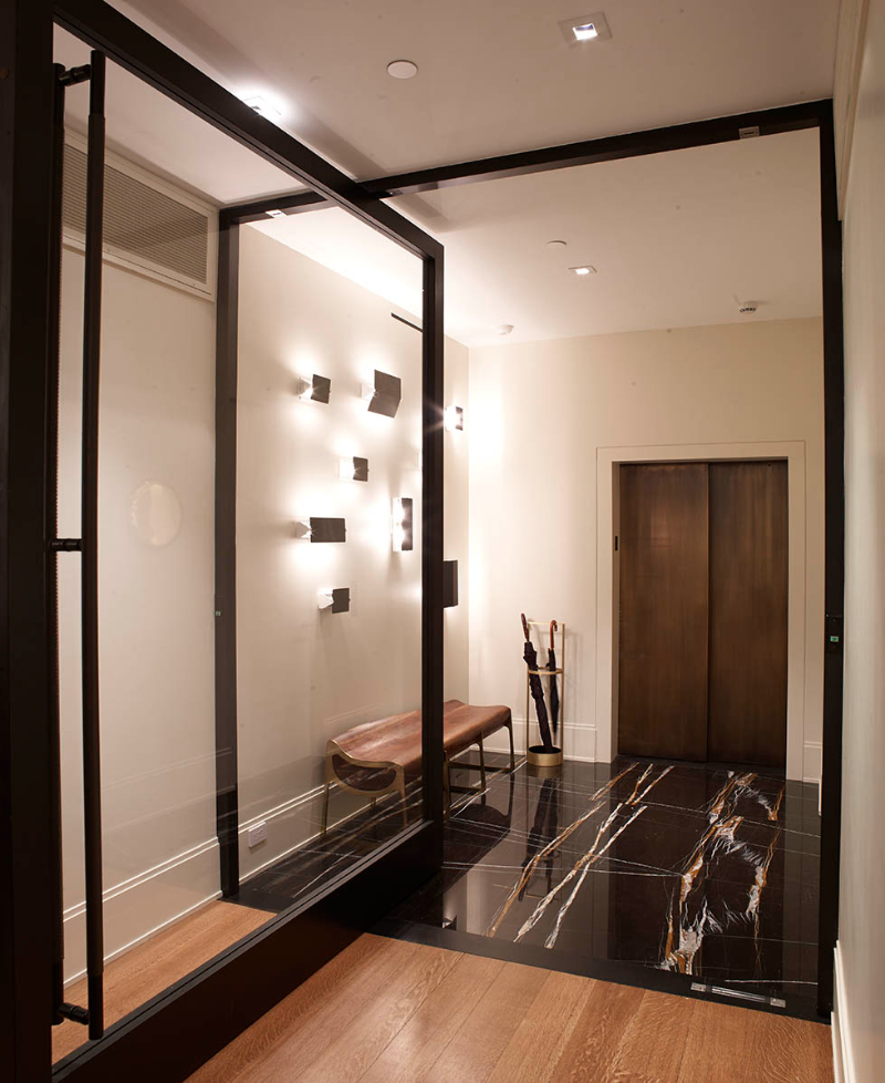 Pembrooke & Ives - A dark marble floor in this entryway, a glass door a bench and some wall lamps.