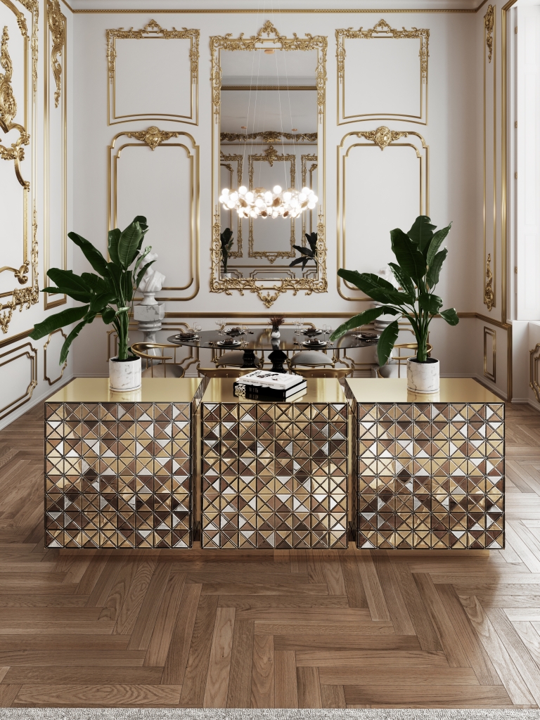 Luxurious dining room featuring the Diamond Sideboard by Boca do Lobo, composed of three mirrored geometric cabinets with gold accents. The space is adorned with ornate gold wall moldings, a grand chandelier, a large decorative mirror, and classical architectural details. Elegant indoor plants and art books add a modern touch to the opulent setting.