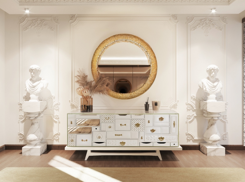 Elegant classical interior featuring the Heritage Sideboard by Boca do Lobo, with a patchwork design in white and gold drawers. A large round gold-leaf mirror hangs above it, flanked by two white marble Roman-style bust sculptures. The ornate wall paneling, wooden flooring, and soft lighting enhance the luxurious ambiance.
