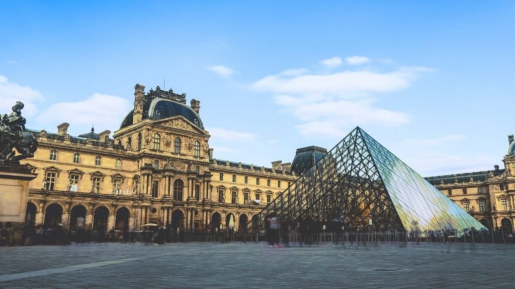 Paris Design Week 2025- Wide-angle view of the historic Louvre Museum building with its classic architecture and the modern glass pyramid entrance under a bright blue sky with scattered clouds.