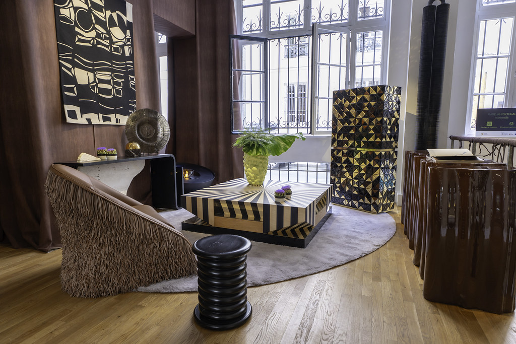 Paris Design Week 2025- Interior view of the Re.MADE IN PORTUGAL exhibition by Nini Andrade Silva, featuring a contemporary lounge setup with a textured brown fringe armchair, a black sculptural stool, a geometric striped coffee table with a green leafy plant centerpiece, and a glossy black and gold patterned cabinet, all set against large windows and brown curtains.
