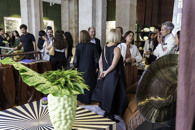 Paris Design Week 2025- People socializing and admiring luxury design pieces at an indoor exhibition, featuring a decorative table with a large green plant in a textured vase in the foreground, elegant furniture, and artistic decor in a well-lit space with stone columns.