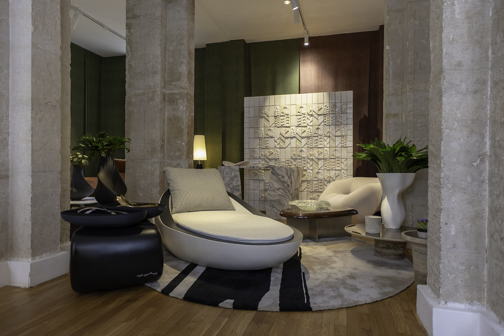 Paris Design Week 2025- Elegant interior scene from the Re.MADE IN PORTUGAL exhibition by Nini Andrade Silva, featuring a modern white chaise lounge with a striped cushion, a soft rounded armchair, black and beige side tables, green plants in white vases, and a textured wall art installation, set between tall stone columns and on a round patterned rug.