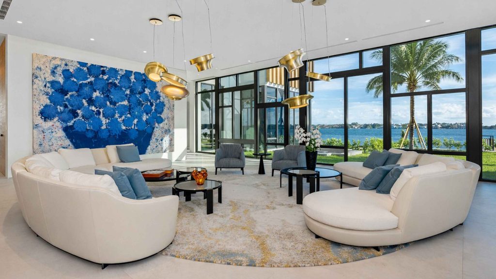 Bright modern living room with large windows, round beige sectional sofa, and a large blue wall art piece.
