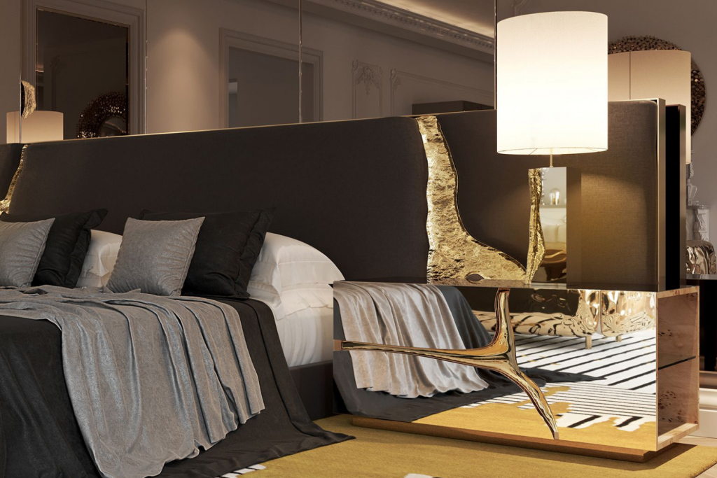 Luxurious bedroom with layered bedding in neutral tones and a mirrored nightstand featuring sculptural gold details, topped by a large white table lamp.