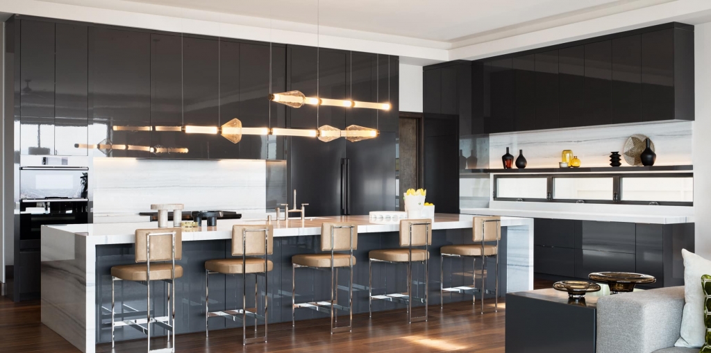 A luxurious modern kitchen featuring sleek, glossy black cabinetry, a large white marble island with tan leather bar stools, and a contemporary linear pendant light fixture.