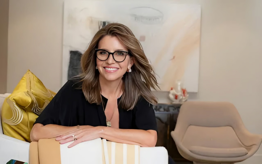 Portrait of a smiling woman with dark-rimmed glasses and brown hair, leaning on the back of a light-colored armchair decorated with yellow pillows, with abstract art hanging on the wall behind her.