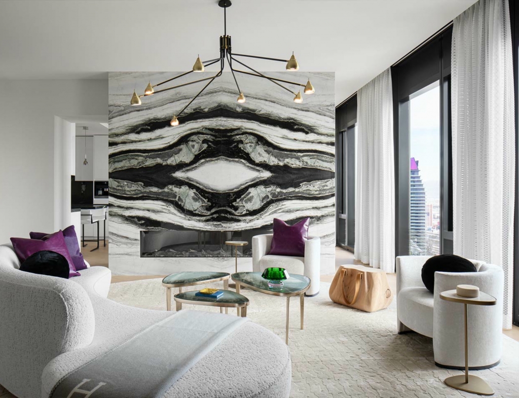 A high-end living room showcasing a dramatic book-matched black and white marble fireplace wall, furnished with curved white textured sofas, purple velvet accent pillows, and organic-shaped nesting tables by floor-to-ceiling windows.