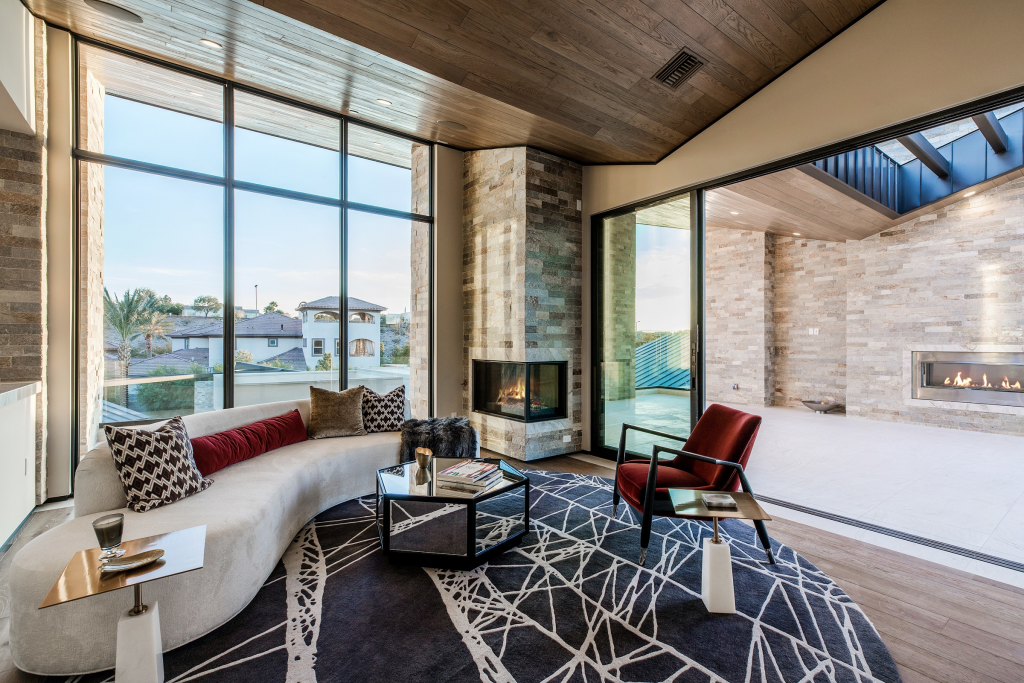 A modern living space with a wood-paneled ceiling, featuring a curved white sofa on a black and white geometric rug, a mirrored hexagonal coffee table, a red armchair, and a stone corner fireplace next to large sliding glass doors.