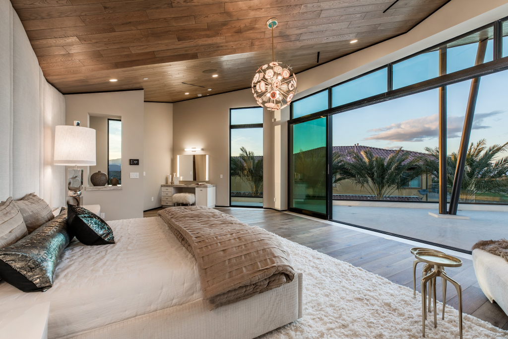 A spacious, upscale bedroom with a wood-paneled ceiling, a plush white area rug under a large bed, a unique round chandelier, and massive sliding glass doors that open to a balcony overlooking palm trees.