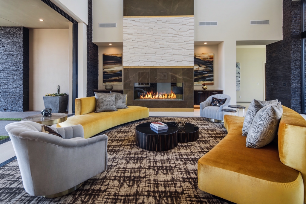 A contemporary living area centered around a tall fireplace with white textured and dark marble accents, symmetrically furnished with two curved yellow velvet sofas, two grey armchairs, and round black coffee tables on a brown patterned rug.