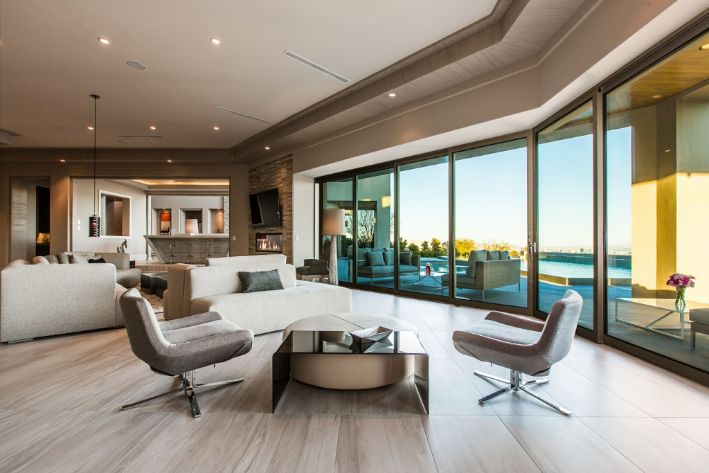 A bright, expansive living room with light tile flooring, featuring a curved light-colored sofa, two grey modern swivel chairs, and large glass walls that slide open to an outdoor patio and pool area.