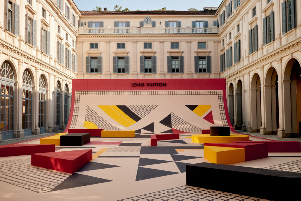 An outdoor courtyard of a historic building is transformed into a modern art and seating area. The ground and a large curved backdrop are decorated with geometric patterns in black, yellow, red, and beige, featuring the word 'LOUIS VUITTON' in red. Large red, yellow, and black geometric-shaped blocks serve as benches. The existing building architecture, with arched windows and light stone, contrasts with the contemporary installation. Objets Nomades