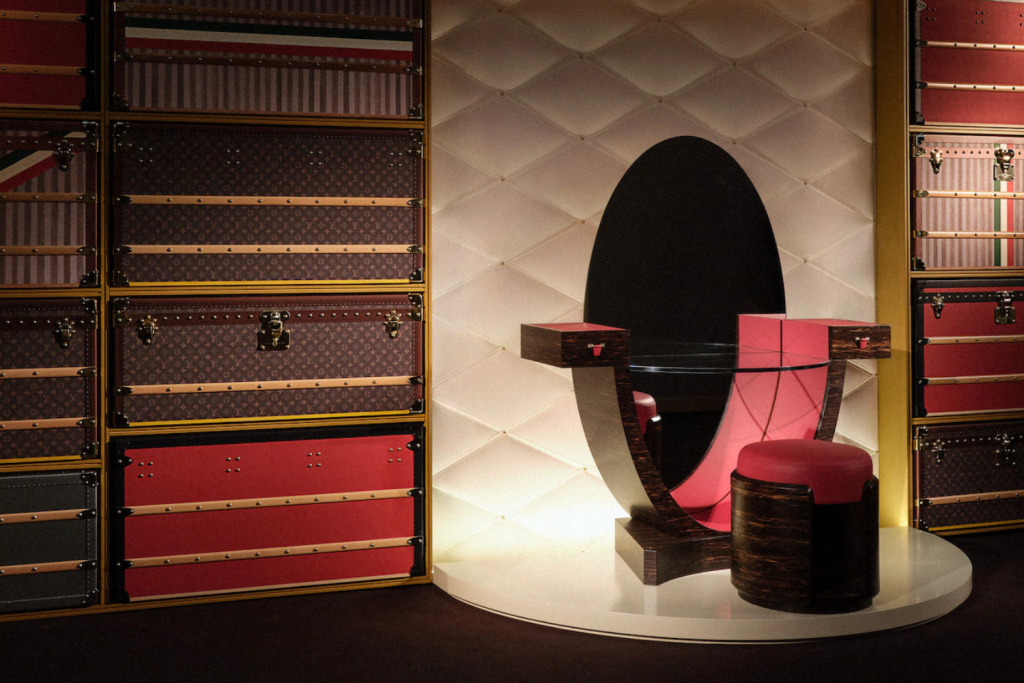 Display of stacked LV trunks surrounding a modern dark-wood vanity and red stool. Objets Nomades