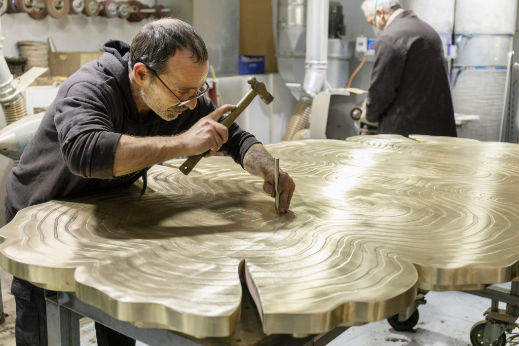 A craftsman in a workshop using a hammer and chisel to detail a large, gold-toned metal table with a tree-ring pattern, Objets Nomades